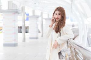 schöne junge asiatische berufstätige frau im weißen anzug lächelt und hält ein smartphone in ihren händen an einem glücklichen arbeitstag mit geschäftsgebäuden und draußen stadt im hintergrund. foto