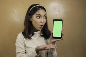 Schönes asiatisches Mädchen ist schockiert, zeigt grünen Bildschirm im Smartphone mit weißem Hemd foto