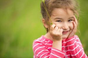 Kleines Mädchen in einem korallenrot gestreiften T-Shirt auf grünem Hintergrund auf einem Feld hält ihr Gesicht in den Händen und lächelt schlau. kindertag, glückliches kind, umwelt- und naturschutz, insektenschutz foto