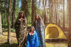 Touristenfamilie aus Vater, Mutter und kleiner Tochter posiert in Schlafsäcken in der Nähe eines Zeltes. Familienerholung im Freien, Inlandstourismus, Camping, Wanderausrüstung. verpuppt wie Raupen-Humor foto