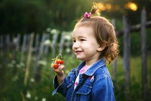 ein kleines mädchen von 2 jahren im dorf hält eine reife erdbeere in ihren händen und lächelt verschmitzt. Vitamine, Sommerzeit. Internationaler Kindertag. Natur, Freiluft foto