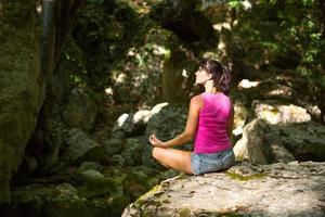Eine Frau sitzt in einem Lotusfeld auf einem großen Felsen zwischen den Felsen im Freien und meditiert, genießt die Einheit mit der Natur, lauscht der Stille und den Geräuschen des Waldes. Ökologie foto