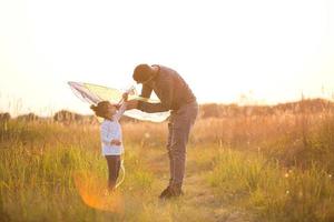 Papa hilft seiner Tochter, im Sommer bei Sonnenuntergang einen Drachen auf einem Feld zu steigen. Familienunterhaltung im Freien, Vatertag, Kindertag. ländliche Gebiete, Unterstützung, gegenseitige Hilfe. orangefarbenes Licht der Sonne foto