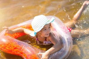 mädchen in einem hut, der im fluss schwimmt, mit einem transparenten aufblasbaren kreis in form eines herzens mit orangefarbenen federn im meer mit sandboden. Strandurlaub, Schwimmen, Bräunen, Sonnencreme. foto