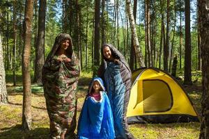 Touristenfamilie aus Vater, Mutter und kleiner Tochter posiert in Schlafsäcken in der Nähe eines Zeltes. Familienerholung im Freien, Inlandstourismus, Camping, Wanderausrüstung. verpuppt wie Raupen-Humor foto