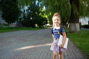 ein kleines mädchen mit kaukasischem aussehen in einer schuluniform mit einem rucksack und dem buch und einer reihe von markierungen. zurück zur Schule. Grundschule, Entwicklung von Aktivitäten für Kinder im Vorschulalter. Platz für Text foto