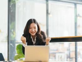 Aufgeregte asiatische Frau sitzt am Schreibtisch und fühlt sich euphorisch, wenn sie Online-Lotterie gewinnt, glückliche schwarze Frau, die überglücklich ist, bekommt Post auf Laptop, die bei der Arbeit befördert wird, gemischtrassiges Mädchen, das erstaunt gute Nachrichten am Computer liest foto