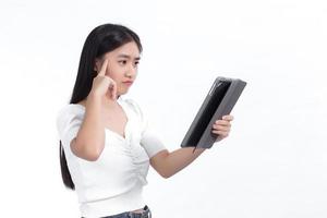 asiatische frau in einem weißen hemd schaut während einer telefonkonferenz auf ein tablet in ihrer hand. vor dem weißen Hintergrund dachte sie glücklich über etwas nach. foto