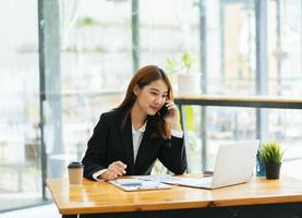 asiatische frau in lässiger kleidung ist glücklich und fröhlich, während sie mit ihrem smartphone kommuniziert und in einem modernen büro arbeitet. foto
