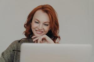 Erfolgreiche Studentin mit rotbraunem Haar und freundlichem Lächeln sieht sich ein interessantes Tutorial-Video oder Webinar an, verwendet einen Laptop für E-Learning, liest Informationen online, hält die Hand unter dem Kinn foto