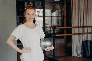 positiver Fitnesstrainer mit Ingwerhaar, der mit einem kleinen silbernen Fitball in der Hand posiert foto