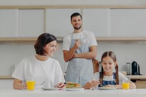 Glückliche Mutter spricht mit Tochter beim Frühstück. vater steht hinter, zubereitetes leckeres gericht für die familie. freundliche Familienmitglieder treffen sich am Wochenende in der Küche, genießen nette Gespräche foto