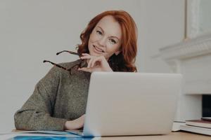 Foto einer fröhlichen rothaarigen Studentin recherchiert für Hausaufgaben, sitzt im Coworking Space im Arbeitszimmer, arbeitet am Laptop, nutzt das Internet. freiberuflicher Webdesigner erstellt Design für Kunden