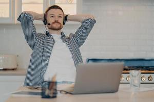 nachdenklicher bärtiger männlicher freiberufler sitzt in entspannter pose macht pause nach der online-arbeit trägt headset mit mikrofon hört bildungs-webinar bekommt fernunterricht nutzt neue computer-app foto