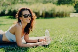 Foto einer fröhlichen, entspannten, brünetten Frau, die auf einer Fitnessmatte posiert, eine trendige Sonnenbrille trägt und Sportkleidung Wasser trinkt, da sie sich durstig fühlt, nachdem Sportübungen, die auf grünem Gras liegen, einen guten sonnigen Tag genießen.