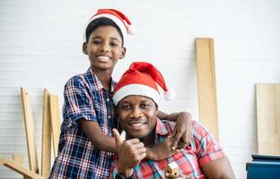 Weihnachten glückliches Kind und Vater. Fröhlicher afroamerikanischer Sohn Tischler, der seinen Vater umarmt, während er sich an den Holztisch lehnt, auf dem verschiedene Arbeitswerkzeuge liegen foto