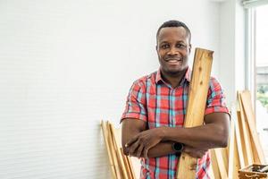 porträt afroamerikaner glücklicher männlicher holzarbeiter oder zimmermann lächelnd, hemd tragend, das arme mit brettholz kreuzt, um diy holzmöbel als hobby herzustellen foto
