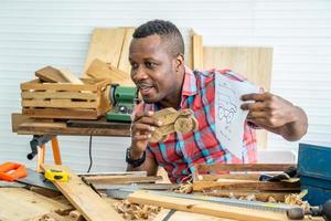 junger afroamerikanischer männlicher Zimmermann, der am Tisch sitzt und Modellautos aus Holz und Zeichenpapier über Tablet zeigt, während er online mit dem Kunden chattet oder online in der Fabrik unterrichtet foto
