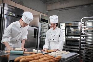 Zwei professionelle asiatische Köche in weißen Kochuniformen und Schürzen kneten Teig und Eier, bereiten Brot, Kekse und frische Backwaren zu und backen in einem Ofen in einer Edelstahlküche des Restaurants. foto