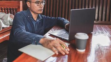 Ein junger Mann mit Brille, der einen Laptop benutzt, der auf einer Tastatur tippt, E-Mails schreibt oder online an einem Computer in seinem Wohnzimmer arbeitet. foto