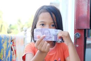 indonesisches kleines Mädchen, das ihren Mund mit Rupiah-Banknoten bedeckt foto