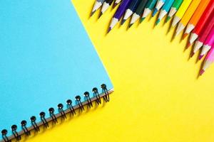 Regenbogenpalette aus Buntstiften mit Spiralblock auf gelbem Hintergrund, verspottet, flach gelegt. Schulanfang, Schülerrekrutierung, Künstler, Zeichenunterricht. Platz kopieren. Schreibwaren für Kreativität foto