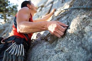 Kletterer im roten T-Shirt klettert auf einen grauen Felsen. Eine starke Hand packte die Führung, selektiver Fokus. Kraft und Ausdauer, Kletterausrüstung Seil, Klettergurt, Chalk, Chalkbag, Karabiner, Hosenträger, Expressschlingen