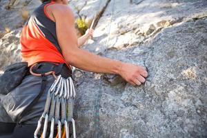 Kletterer im roten T-Shirt klettert auf einen grauen Felsen. Eine starke Hand packte die Führung, selektiver Fokus. Kraft und Ausdauer, Kletterausrüstung Seil, Klettergurt, Chalk, Chalkbag, Karabiner, Hosenträger, Expressschlingen