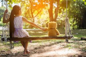 Ein kleines Mädchen und ein Teddybär sitzen auf einer Schaukel. foto