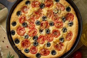Focaccia, Pizza in der Pfanne, italienisches Fladenbrot mit Tomaten, Oliven und Rosmarin. Holztisch