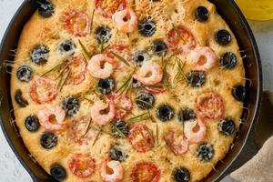 Focaccia, Garnelenpizza in der Pfanne. Nahaufnahme italienisches Fladenbrot mit Tomaten, Oliven und Rosmarin. Draufsicht, weißer konkreter Hintergrund foto