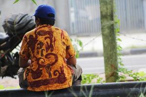 ein mann mittleren alters sitzt am straßenrand in jakarta und trägt indonesische batikkleidung foto