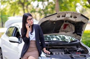 eine junge asiatin ruft ihren servicetechniker an, um ein kaputtes auto am straßenrand zu reparieren foto