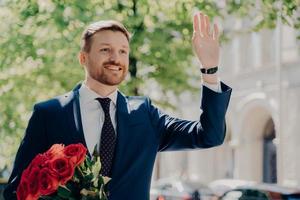 Porträt eines wunderschönen, eleganten Mannes im Smoking mit einem Strauß roter Rosen draußen im Stadtzentrum foto