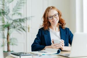 innenaufnahme einer redakteurin arbeitet an einem artikel, verwendet einen laptop und drahtloses internet, überprüft die neuesten nachrichten, hält eine tasse kaffee oder tee, umgeben von einem notizblock zum schreiben von notizen, lächelt glücklich foto