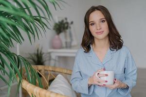 Foto einer hübschen Frau mit ansprechendem Aussehen, gekleidet in lässige Hauskleidung, hält eine weiße Tasse mit aromatischem Kaffee oder Tee, sitzt auf Korbmöbeln im Innenbereich mit Platz für Ihre Werbung