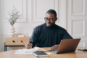 Fokussierter junger afroamerikanischer Geschäftsmann, der während der Arbeit im Büro Notizen in ein Notizbuch aufschreibt