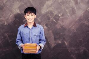 hübscher junger asiatischer Mann, der ein Buch auf dem Grunge-Wand-Smiley hält und in die Kamera schaut foto