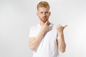 junger lässiger mann, der etwas hinterhältig auf den kopierraum schaut. hübscher junger kaukasischer mann im t-shirt lokalisiert auf weißem hintergrund. foto