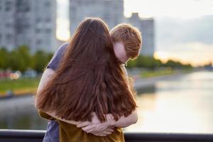 Mädchen mit langem, dickem, dunklem Ohr, das einen rothaarigen Jungen auf der Brücke umarmt, jugendlich Liebe bei Sonnenuntergang foto