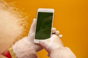 weihnachtsmann, der unter verwendung des smartphones auf gelber texturhintergrundoberfläche arbeitet. Nahaufnahme von oben auf die Hände. frohes neues Jahr und frohe Weihnachten foto