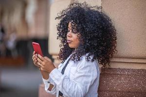 junge schwarze frau mit lockigem haar, die mit dem handy geht. SMS auf der Straße. große Stadt. foto