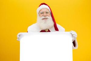 glücklicher weihnachtsmann, der hinter dem leeren schild herausschaut, das auf gelbem hintergrund mit kopienraum isoliert ist foto