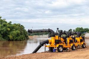 Eine große gelbe Pumpe mit schwarzen Rohren ist auf dem Boden des Flussufers montiert, um das Pumpen in der Nähe eines Damms vorzubereiten, einer Betonsäulenwand, um Erosion zu verhindern. foto