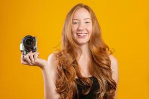 Foto einer entzückenden rothaarigen Frau in schwarzer Kleidung, die eine Retro-Vintage-Kamera hält und ein Foto isoliert über gelbem Hintergrund macht. kopierraum nachbauen. Halten Sie Retro-Vintage-Fotokamera.