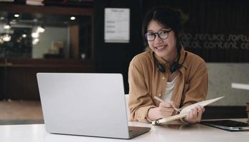 glückliche asiatische frau mit headset macht sich notizen, spricht per konferenzgespräch und macht video-chat auf laptop im büro, lächelnder chinesischer support service agent schreibt informationen konsultieren kunden foto