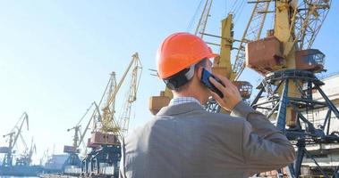 junger männlicher arbeiter des seehafens im helm, frachtmanager im anzug und halmet arbeitet im freien, kräne und meereshintergrund foto