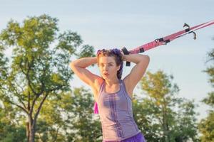 schöne fitte frau in rosa und lila sportbekleidung morgens training im outdoor-fitnessstudio, übungen mit aufhängungsgurten im park foto