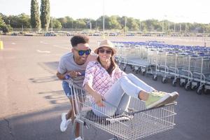 Fröhliches junges Paar, das auf einem leeren Parkplatz in einem Einkaufszentrum fährt, ein Hipster-Freund hat eine gute Zeit beim Einkaufen, ein verliebtes Paar, das auf einem Einkaufswagen reitet foto