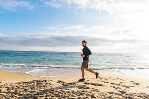 Passen Sie das männliche Läufertraining am Sommerstrand an und hören Sie Musik gegen den wunderschönen Himmel und das Meer foto